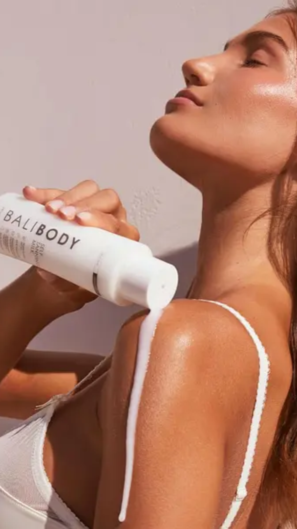 Woman applying sunscreen cream to her shoulder with a 'Bali Body' bottle.