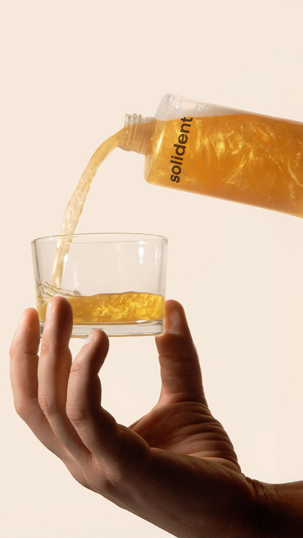 Hand pouring a yellow liquid from a bottle labeled 'solident' into a glass against a beige background.