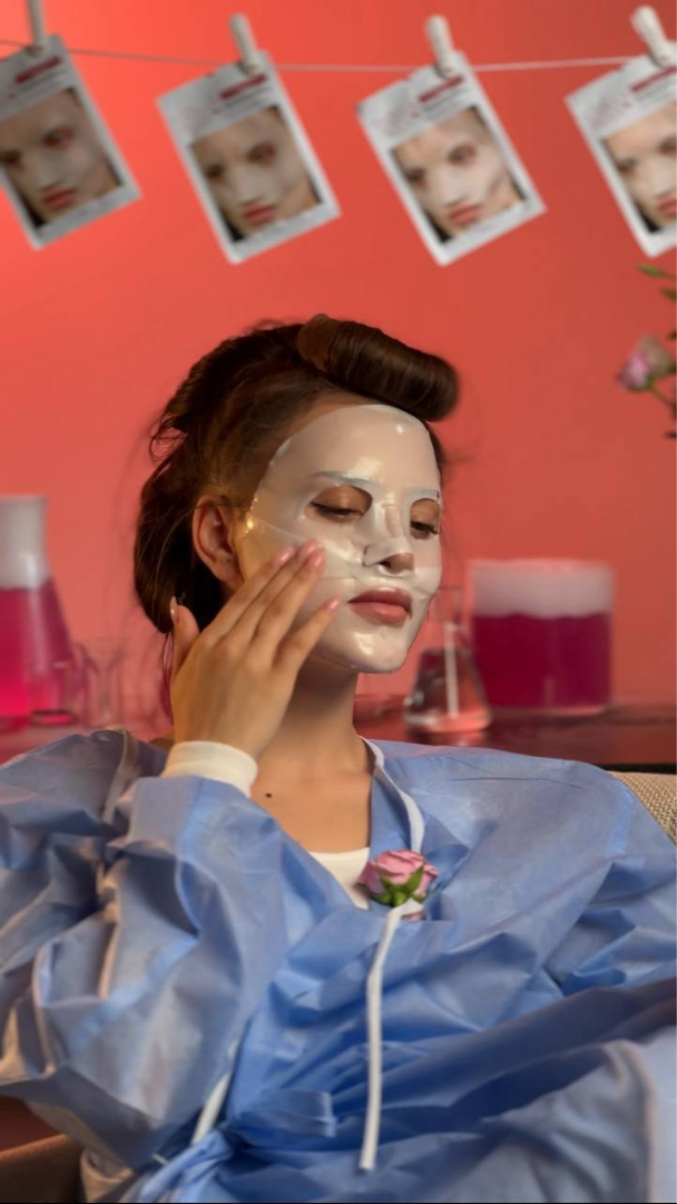 Person applying a face mask with a pink background and posters on a string.