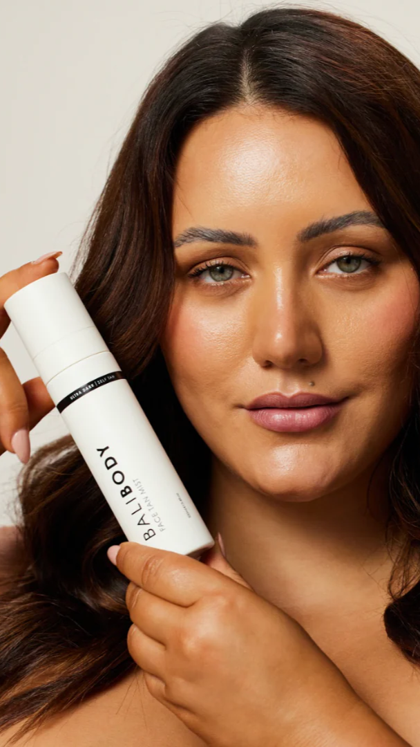 Woman holding a white spray bottle labeled 'Ballyhoo' against a neutral background
