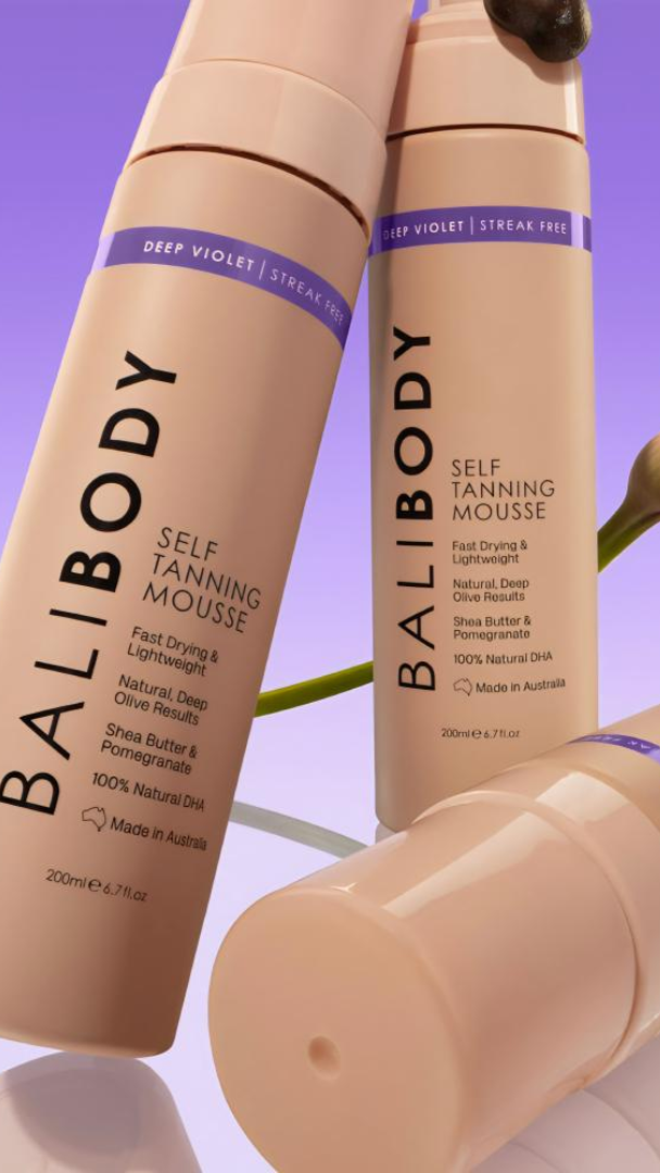 Three bottles of Bali Body self-tanning mousse on a purple background with a flower.