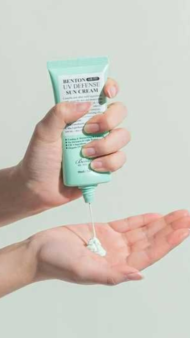 Hand holding a tube of Benton UV Defense Sun Cream with cream being squeezed onto the palm against a light gray background