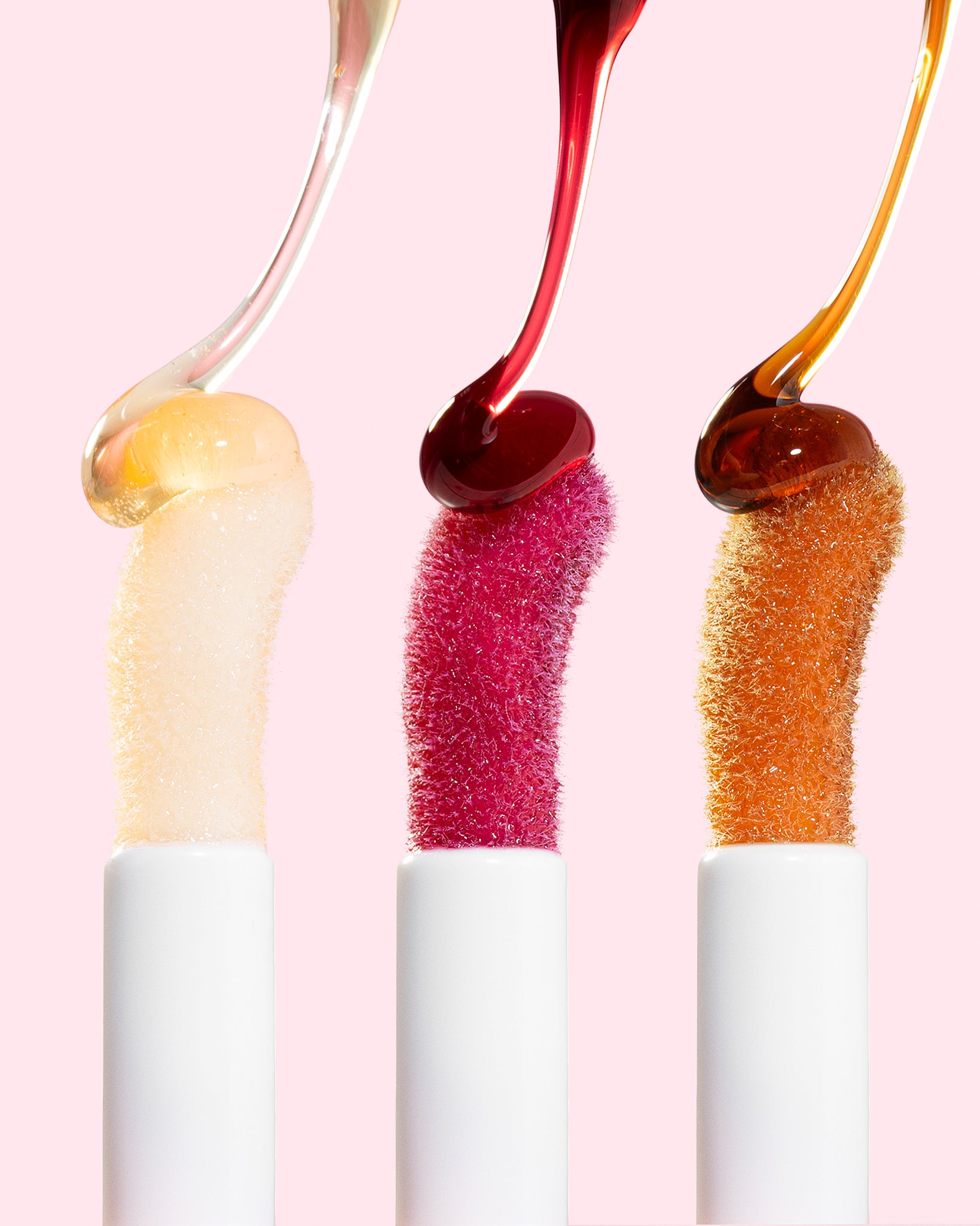 Three lip glosses with colorful liquids dripping from them on a pink background
